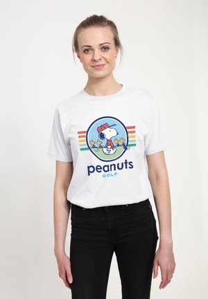 Lachende vrouw die een wit T-shirt draagt met een Snoopy en Woodstock golfafbeelding en de tekst "peanuts GOLF", gecombineerd met een zwarte broek.