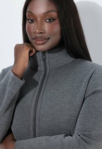 Veste grise zippée avec un col haut, une texture douce et des manches structurées. Comprend une fermeture éclair centrale avec un détail en tissu contrastant.