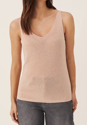 Kvinde iført en lyserød strikket ærmeløs top med V-hals, parret med mørkegrå denimjeans, stående mod en ensfarvet baggrund.