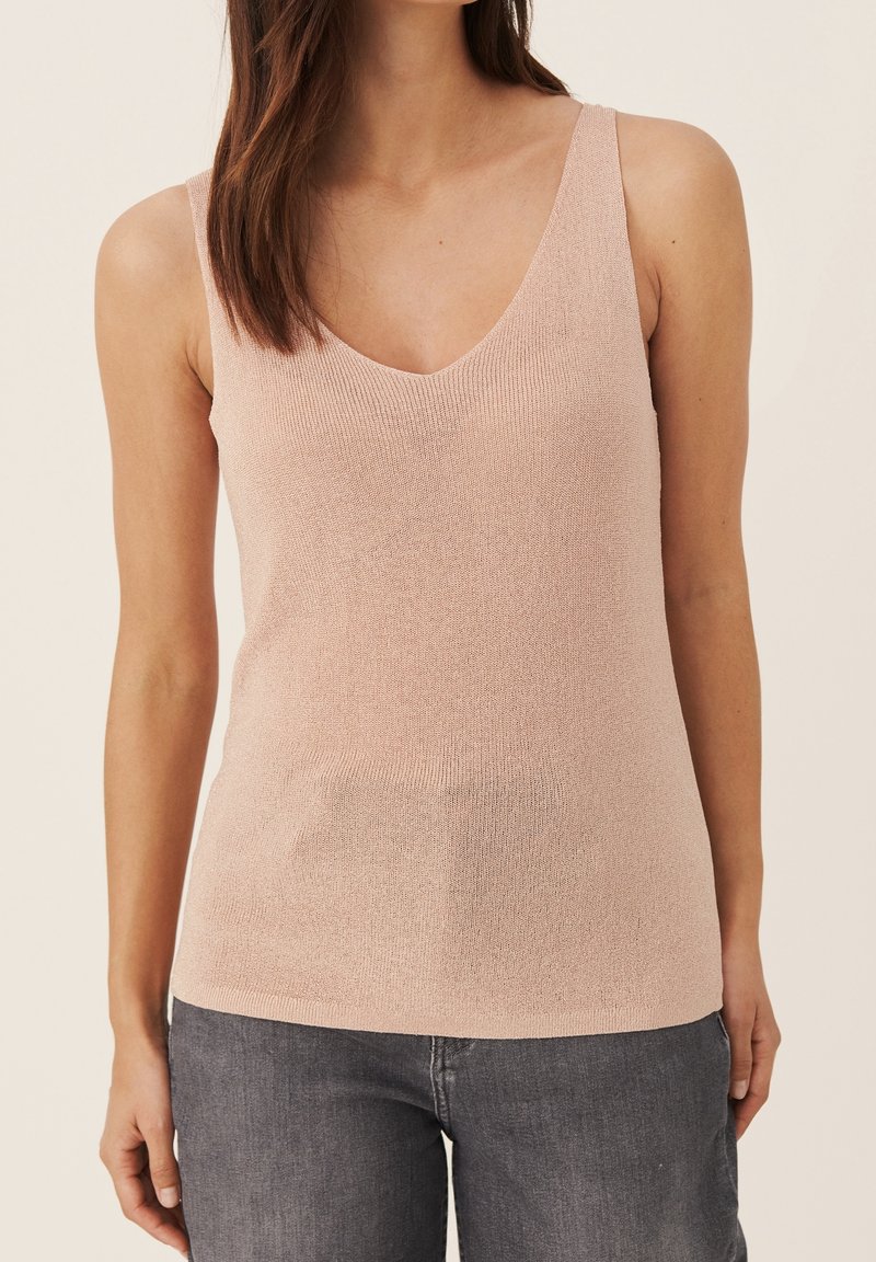 Femme portant un haut sans manches en tricot rose clair avec un col en V, associé à un jean en denim gris foncé, debout devant un fond uni.