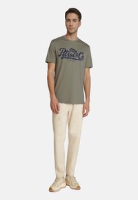 Oliven grøn t-shirt med "Petrol & Co" grafik i marineblå, korte ærmer, parret med beige bukser og lyse sneakers. Glat tekstur.