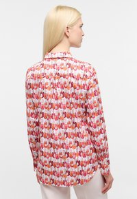 Camicia a maniche lunghe con colletto in tessuto leggero, decorata con un motivo ripetuto di boccette di smalto per unghie rosa e arancione su sfondo bianco.