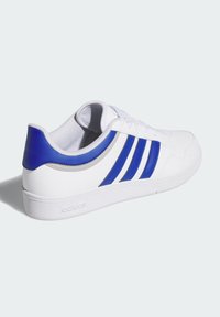 Zapatilla deportiva blanca con tres franjas azules, parte superior de cuero, acento en el talón texturizado en azul y suela de goma. Punta reforzada.