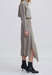 Frau in taupefarbenem, cropped Blazer und passendem Midi-Rock mit geripptem Einsatz, seitlichem Schlitz und schwarzen, hohen Stiefeletten, steht seitlich.