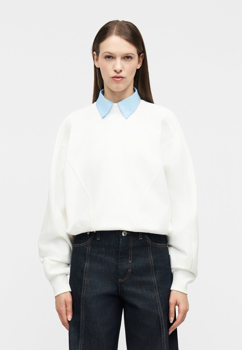 Γυναίκα με ίσια καστανά μαλλιά, που φορά λευκό φούτερ πάνω από ένα ανοιχτό μπλε πουκάμισο με γιακά και σκούρο τζιν ψηλόμεσο παντελόνι, στέκεται μπροστά σε λευκό φόντο.