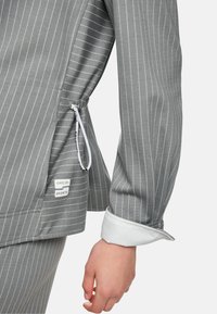 Grauer Nadelstreifenblazer mit Kordelzug am Saum und weißen Manschetten. Enthält ein Etikett mit der Markenbezeichnung "Marc Cain Sports".