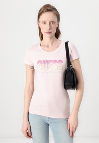 T-shirt rose clair à manches courtes, avec un logo "GUESS" dégradé orné de strass ; associé à un sac à épaule noir.