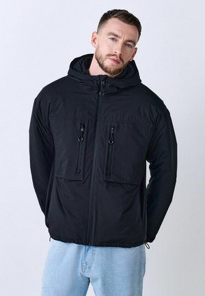 Chaqueta negra con cremallera y capucha, dos bolsillos frontales con cremalleras y una textura suave. Combinada con pantalones de mezclilla azul claro.