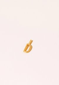 Guldfärgad bokstavspendant med design i elegant utskärning. Slät yta, liten ögla för fästning, mot en ljus bakgrund.