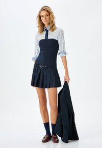 Marineblaues trägerloses Kleid mit gefälteltem Rock, kombiniert mit einem blau-weiß gestreiften Hemd, einer marineblauen Krawatte und dunklen Loden; hält einen dunklen Blazer.