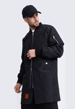 Zwarte lichtgewicht bomberjack met een voorkant met rits, zijzakken, geribbelde manchetten en een oranje label op de zoom. Draagt een zwarte pet.