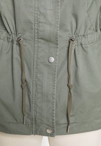 Veste en toile verte avec une taille à cordon de serrage, des fermetures à pression argentées et un tissu texturé avec des détails de couture subtils.