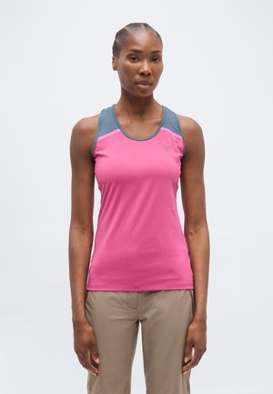 Vrouw draagt een mouwloos roze en grijs sporttopje en beige broek, staand tegen een effen lichte achtergrond.