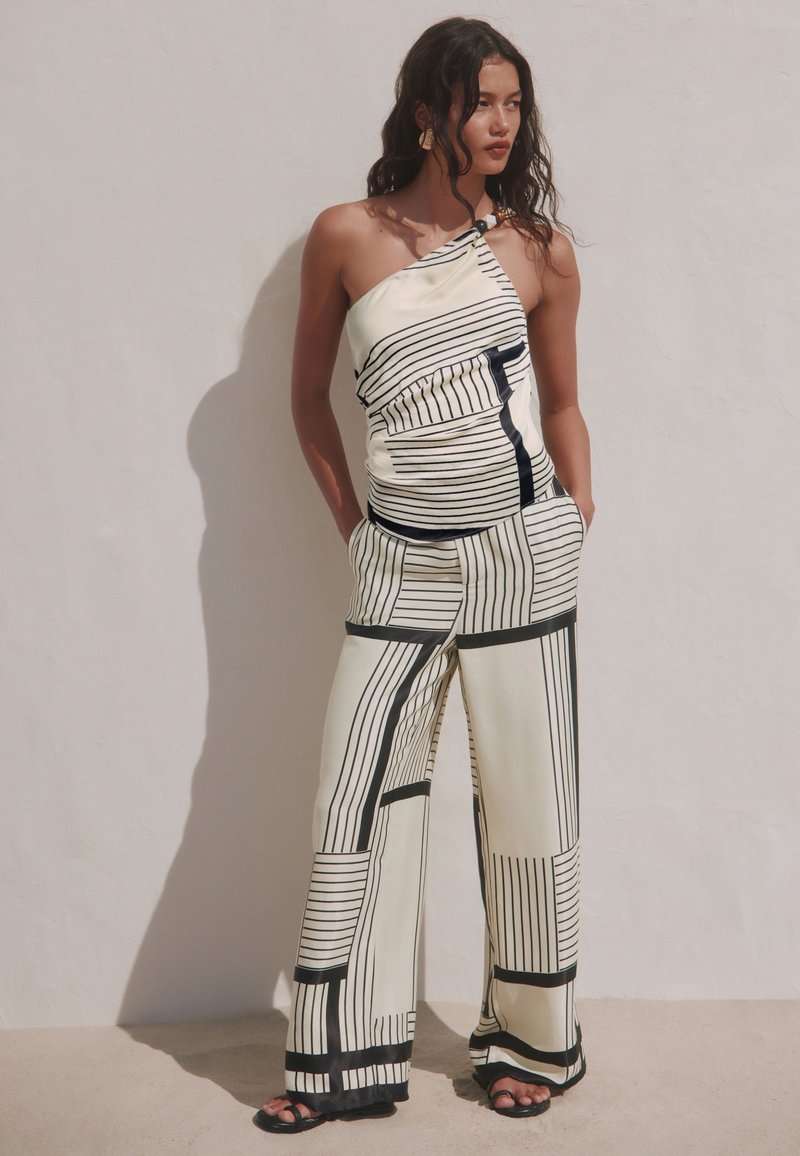 Femme aux cheveux foncés ondulés, portant un haut asymétrique sans manches et un pantalon large à rayures géométriques noires et blanches, debout contre un mur clair.