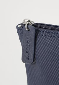 Navy blue textured pouch with silver zipper and Lacoste brand tag. Features a distinctive pattern and durable material. Compact and functional design.