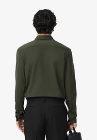 Camicia verde scuro a maniche lunghe con colletto, dotata di polsini con bottoni e taglio dritto. Abbinata a pantaloni neri e a una borsa nera.