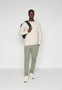 Vaaleanbeige pitkähihainen paita ja oliivinvihreät cargo-housut, varustettuna mustalla repulla. Kengät ovat valkoiset tennarit, joissa on vaaleanbeigejä yksityiskohtia.