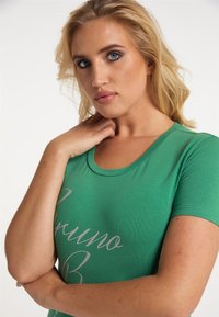 Grön kortärmad T-shirt i mjukt tyg, med en båtringning och tryckt text i ljusgrått över framsidan.