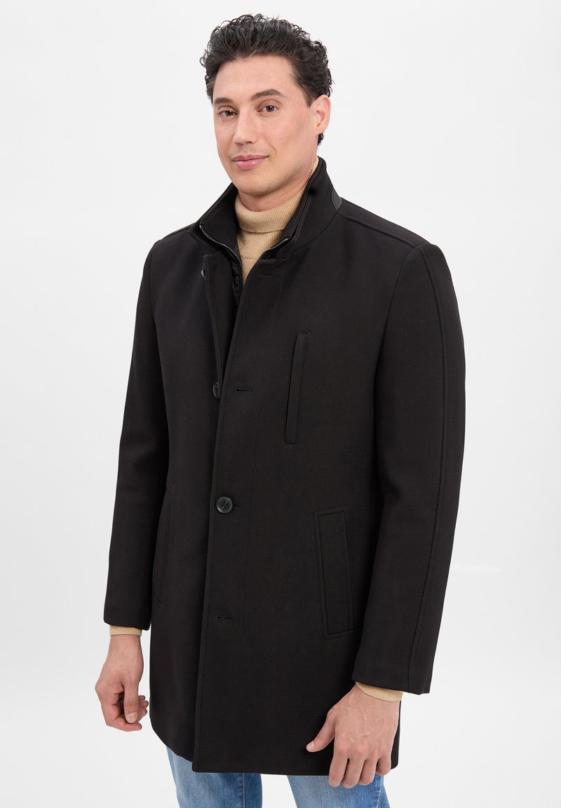 Schwarzer Mantel mit hohem Kragen, vorderer Knopfverschluss und seitlichen Taschen. Hergestellt aus strukturiertem Stoff mit dezenten Musterakzenten.