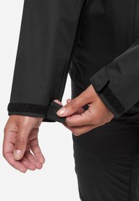 Schwarze isolierte Jacke mit strukturiertem Stoff, der eine Klettverschluss-Manschettenverstellung zeigt. Der Fokus liegt auf einer Hand, die die Manschette greift, mit glatten, subtilen Details.