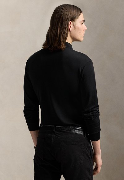 Jeune homme aux cheveux bruns mi-longs, portant un haut noir à manches longues et un pantalon noir, de profil sur un fond neutre.