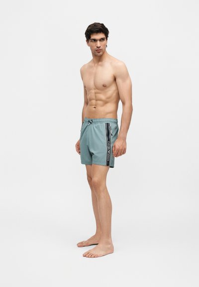 Νεαρός άνδρας στέκεται ξυπόλητος φορώντας ανοιχτογάλανα μαγιό με μαύρη κάθετη ρίγα και τσέπη με φερμουάρ σε λευκό φόντο.