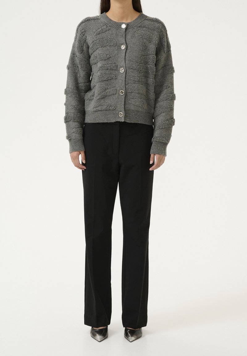 Cardigan grigio a trama con scollatura rotonda, cinque bottoni argentati e polsini a coste, abbinato a pantaloni neri e tacchi a punta.