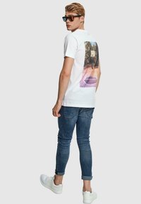 Tricou alb din bumbac cu un grafic colorat pe spate. Poartă blugi de culoare albastru închis și adidași albi. Modelul poartă ochelari de soare.
