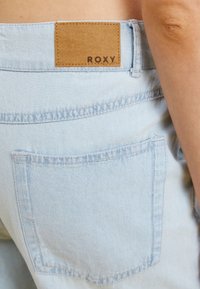 Svijetloplave traperice s tamnom kožnom oznakom označenom "ROXY" na stražnjem struku, imaju jedinstveni stražnji džep i detalje šivanja.