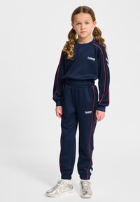 Marinträningsoverall i mjukt material, med röda sidostreck och logodetaljer. Byxorna har ett elastiskt midjeband och är parat med reflekterande sneakers.