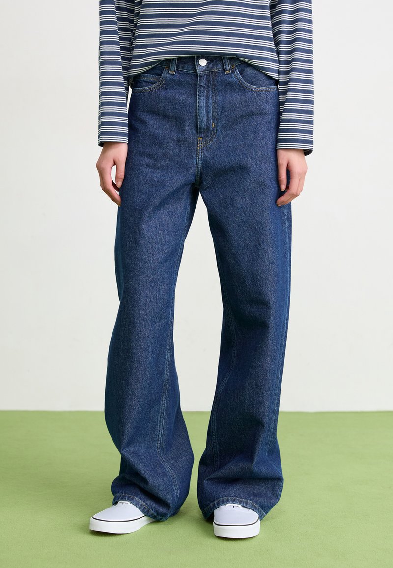 Jeans a gamba larga blu scuro in denim con vita alta, abbinati a una maglietta a maniche lunghe a righe e scarpe slip-on bianche.