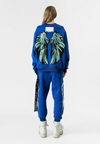 Sudadera azul con un gran diseño de alas verdes y negras en la espalda, combinada con pantalones a juego. Presenta una etiqueta blanca y botas con cremallera.