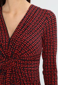 Robe noire avec un décolleté en V, des manches longues et un détail de nœud torsadé à la taille. Motif géométrique rouge sur l'ensemble. Texture de tissu douce.