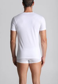 Uomo visto da dietro che indossa una maglietta bianca aderente a maniche corte e slip bianchi coordinati su uno sfondo grigio semplice.