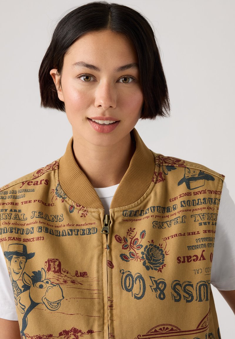 Tan zip-up vest med trykte grafikker, der inkluderer tekst og illustrationer, ribbet krave og struktureret stof. Bæres over en hvid skjorte.