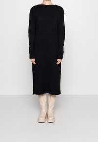 Černé pletené šaty s dlouhým rukávem a bočními rozparky, kombinované s béžovými kozačkami po kolena. Látka vypadá měkce a má texturu.