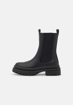 Botte Chelsea en cuir noir avec semelle en caoutchouc épaisse, panneaux latéraux élastiques, languettes de tirage, et bout arrondi sur fond blanc.