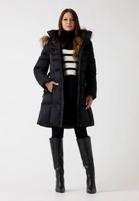 Cappotto smanicato nero con cappuccio di pelliccia, chiusura frontale con zip e tasche laterali, indossato sopra un maglione a righe. Stivali neri alti completano il look.