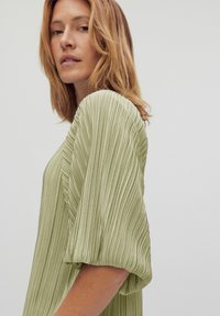 Vaaleanvihreä, laskostettu yläosa, jossa on lyhyet, puffimaiset hihat. Sileä kangas ja vartalonmyötäinen siluetti, joka korostaa kangasta vaakasuoria viivoja.