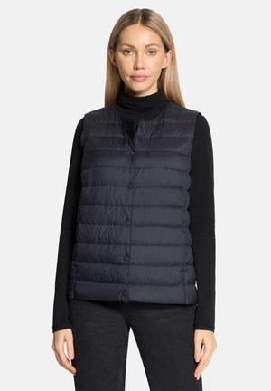 Kvinde iført en sort langærmet top, mørk vatteret vest med trykknapper og mørke bukser mod en ensartet lys baggrund.