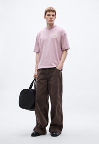 Svetloružové bavlnené tričko s krátkymi rukávmi a vyšívaným detailom, kombinované s hnedými širokými nohavicami a čiernou okrúhlou taškou typu duffel.