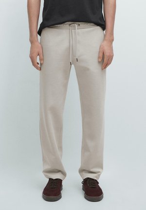 Uomo che indossa pantaloni della tuta beige con coulisse e scarpe da ginnastica rosso scuro, in piedi davanti a uno sfondo chiaro e semplice.