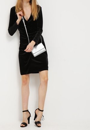 Žena v černých sametových šatech s dlouhým rukávem drží stříbrnou kabelku přes rameno, má na sobě černé sandály na vysokém podpatku a stojí u bílé zdi.