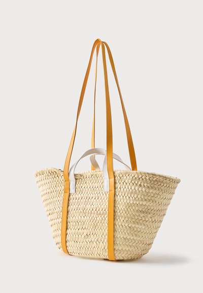 Πλεκτή τσάντα από ψάθα με μακριά λουριά ώμου από καφέ δέρμα και κοντά λευκά χερούλια, τοποθετημένη σε απλό ανοιχτό φόντο.