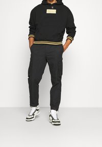 Sudadera negra con etiqueta dorada y puños de canalé, combinada con pantalones cargo negros con bolsillos laterales. Llevada con zapatillas verdes y blancas.