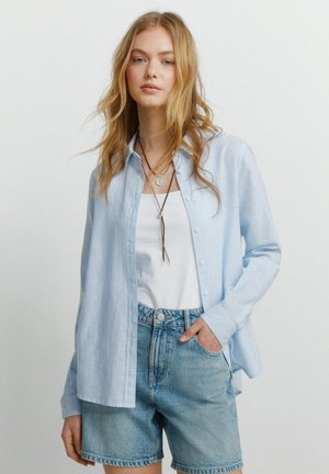 Młoda kobieta z długimi blond włosami, ubrana w jasnoniebieską koszulę na guziki na białym topie i dżinsowe szorty, z ręką w kieszeni.