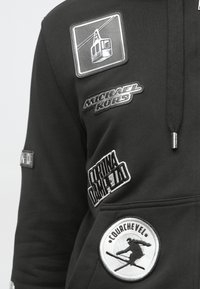 Μαύρο φούτερ με πολλαπλές επιδιορθώσεις: μία με σχέδιο λιφτ σκι, λογότυπο "Michael Kors" και δύο άλλες κεντημένες ετικέτες.