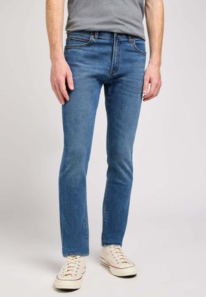 Jeans slim fit da uomo in un lavaggio blu medio, con design a cinque tasche, chiusura con zip e dettagli di cucitura discreti. Abbinati a sneakers bianche.