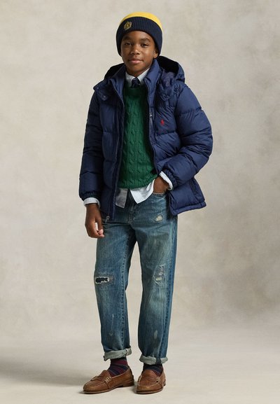 Băiat purtând o geacă puf albastru închis, pulover verde, cămașă albă, cravată, blugi rupți suflecați, mocasini maro și o căciulă albastru închis-galben, stând cu o mână în buzunar.