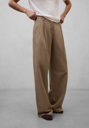 Person iført beige bukser med vide ben og en hvid kortærmet skjorte, justerer linningen, står på en grå gulv mod en ensfarvet væg.